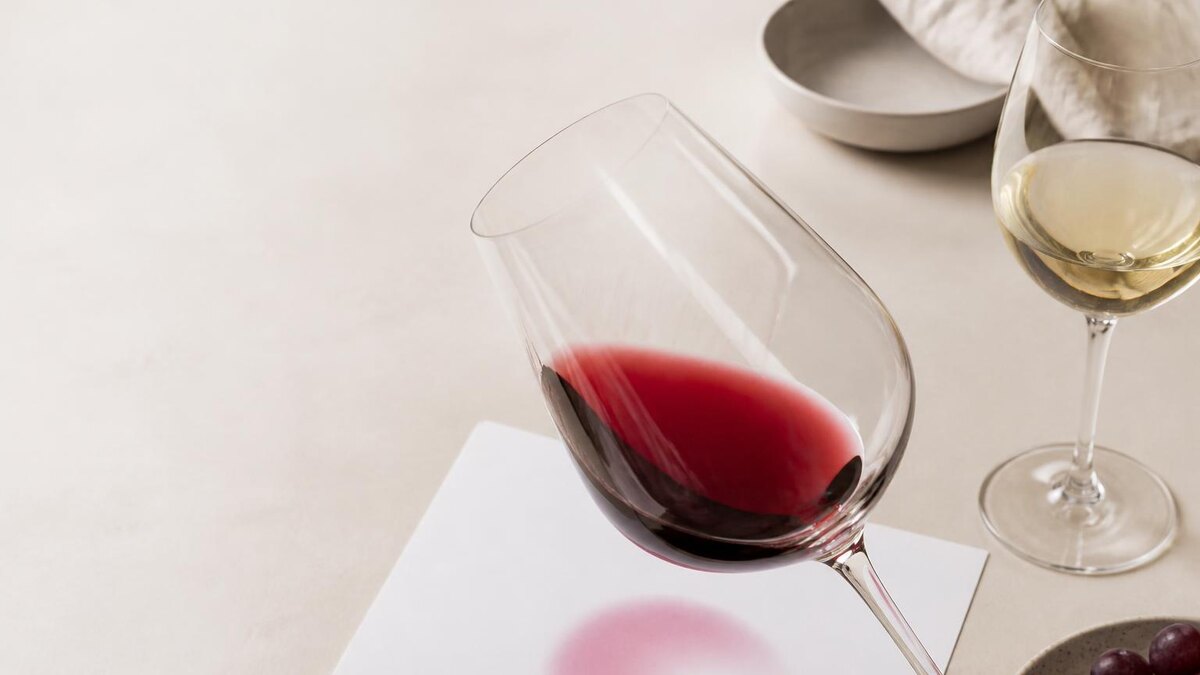 Ruhige, realistische Weinverkostungsszene im August bei neutralem Tageslicht: ein farbloses Weinglas mit Rotwein leicht über einem weißen Bl
